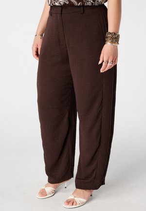 Vrouw draagt losse, donkerbruine gestructureerde broek, witte sandalen met bandjes en hakken, en armbanden aan haar rechterpols.