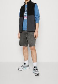 Svart och grå fleeceväst med hög krage, blå sweatshirt, grå shorts och silverfärgade löparskor med svarta och turkosa detaljer.