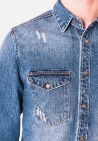 Camicia in denim di blu medio, con colletto a punta, tasca frontale e dettagli usurati con bottoni a pressione in metallo. Texture liscia.