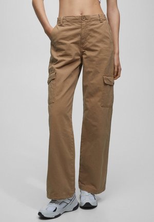 Person iført brune cargobukser og hvide sneakers, stående med hænderne i lommerne mod en ensfarvet lys baggrund.