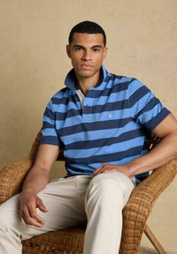 Homme assis dans une chaise en osier portant un polo à rayures bleu et marine et un pantalon beige clair sur un fond neutre.