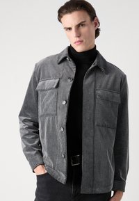 Gray corduroy jacket with vertical stripes, featuring two chest pockets, button closure, and a collared neckline. Worn over a black turtleneck.