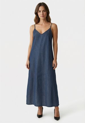 Mujer de pie contra un fondo blanco, vistiendo un vestido largo hasta el tobillo de mezclilla azul oscuro sin mangas con tirantes finos y tacones altos negros.