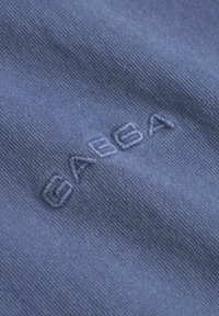 Modrá bavlnená tkanina s hladkým povrchom, na ktorej sa nachádza vyšívané logo "GABBA" v mierne tmavšej modrej niťi.