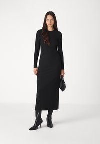 Black ribbed long-sleeve dress with a round neckline, fitted silhouette, and a midi length; paired with black high-heeled boots and a clutch.