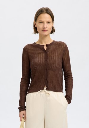 Cardigan marron à côtes avec un col rond, des manches longues et une fermeture par boutons. Porté par-dessus un pantalon clair et ample.