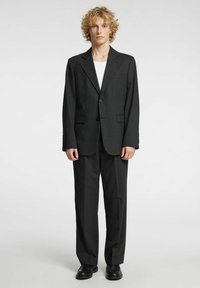 Completo nero con blazer strutturato e pantaloni a gamba dritta. Presenta un risvolto a incavo, due tasche frontali e tessuto liscio. Scarpe nere completano il look.