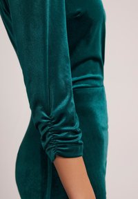 Zielona sukienka z aksamitu o dopasowanej sylwetce, z marszczeniami przy nadgarstkach oraz gładkiej fakturze, która subtelnie odbija światło.