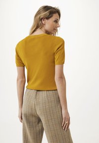 Kvinna med blont hår som bär en senapsgul kortärmad topp och högmidjade beige rutiga byxor, ser över sin axel.