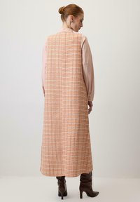 Long, sleeveless dress in orange woven fabric with a checked pattern, paired with a light pink shirt and brown heeled boots.