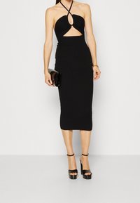 Czarna, prążkowana sukienka midi z dekoltem na halter, wycięciem z przodu i detalem w kształcie dziurki. Modelka trzyma czarną kopertówkę i ma na sobie czarne platformy.