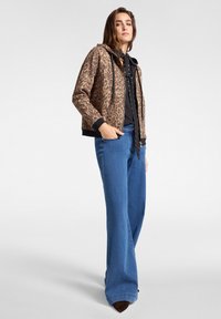 Giovane donna con giacca maculata, blouse nera con dettaglio a fiocco, jeans a gamba larga blu e scarpe a punta marroni, in piedi con una mano in tasca.