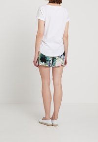 T-shirt blanc en coton à manches courtes, associée à un short coloré à motifs floraux avec des nuances de vert et de rose, et des chaussures blanches sans lacets.