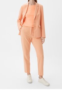 Costume pêche comprenant un blazer ajusté à deux boutons et un pantalon fuselé assorti. Porté avec des baskets claires. Tissu lisse.