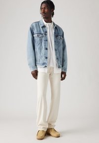 Hombre de pie vistiendo una chaqueta de mezclilla azul clara, una camisa blanca, pantalones color crema y zapatos de ante beige sobre un fondo liso.