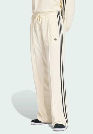 Cremefarbene Adidas Trainingshose mit schwarzen drei Streifen an den Seiten, einem Tunnelzug in der Taille und schwarzen Slip-On-Schuhen.