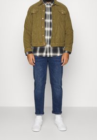 Olivenfarbene Cordjacke mit zwei Brusttaschen, kombiniert mit einem schwarz-weißen Karoshirt, blauen Jeans und weißen Sneakers.