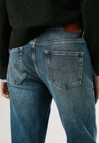 Tummansinisiä korkeavyötäröisiä farkkuja, joissa on kaksi takataskua ja näkyvät ompelukoristeet. Materiaali vaikuttaa pehmeältä denimiltä.