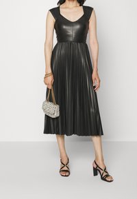 Robe noire plissée avec un corsage en cuir ajusté, des manches courtes et une jupe fluide. Associée à un petit sac à main texturé et des talons avec lanières.