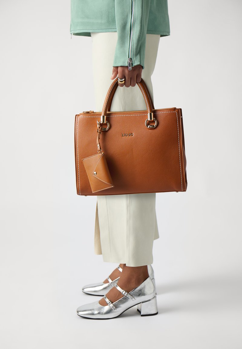 Borsa a tracolla in pelle marrone con texture liscia, hardware dorato e un'etichetta removibile. Sono visibili anche pantaloni bianchi e scarpe con tacco a blocco argentate.