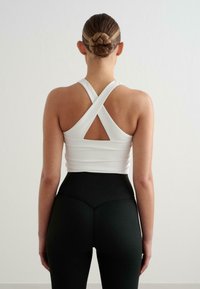 Fehér sportos tank top, kereszt hátú dizájnnal, kivágott részlettel. Magas derekú fekete leggingssel párosítva. Sima textúra.