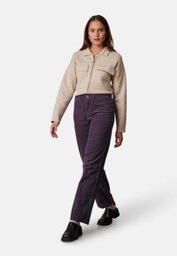 Beige zip-up jas met twee voorzakken, gecombineerd met paarse corduroy broek. Model draagt zwarte platform schoenen en loopt tegen een witte achtergrond.