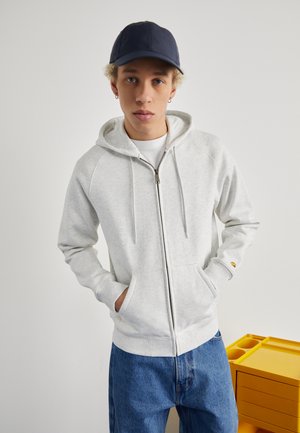Light gray zip-up hoodie with drawstrings, kangaroo pockets, and ribbed cuffs. Paired with loose blue jeans and a navy cap.