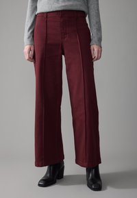 Persona que lleva pantalones burdeos de cintura alta y pierna ancha, una camiseta gris de manga larga y botines negros de tacón, de pie frente a un fondo gris.