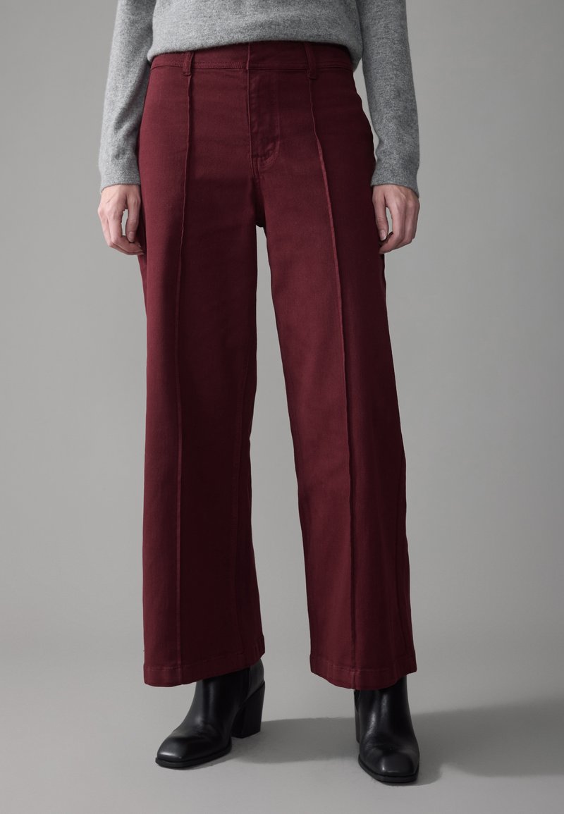 Persona que lleva pantalones burdeos de cintura alta y pierna ancha, una camiseta gris de manga larga y botines negros de tacón, de pie frente a un fondo gris.