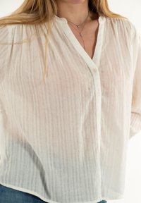 Camisa blanca de botones con textura, rayas verticales y escote en V. Presenta un corte holgado y una tela suave, con mangas largas ligeramente fruncidas.