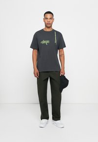 Mørkegrå t-shirt med grøn Nike-logo, parret med olivengrønne cargo-bukser og hvide sneakers. Sort kasket holdt i højre hånd.