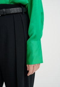 Camicia verde in satin a maniche lunghe abbinata a pantaloni neri a vita alta. La camicia presenta una texture liscia e polsini strutturati. Cintura nera strutturata inclusa.