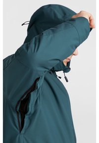 Chaqueta impermeable en color teal con capucha, que cuenta con bolsillos con cremallera, puños elásticos y una textura suave y mate. Corte ajustado y atlético.