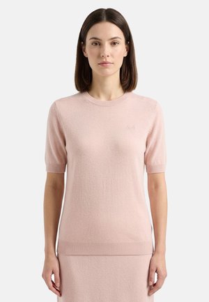 Donna con capelli castani fino alle spalle che indossa un top lavorato a maglia rosa chiaro a maniche corte e gonna abbinata su uno sfondo semplice.