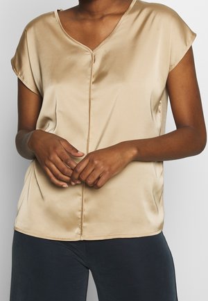 Personne portant un chemisier en satin beige à manches courtes avec un col en V et un pantalon noir, mains jointes à la taille.