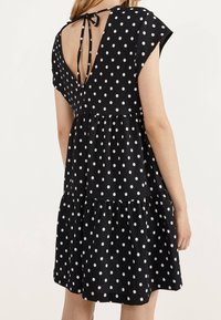 Kvinna klädd i en svart klänning med vita prickar, med en djup V-rygg och knytning högst upp på ryggen.