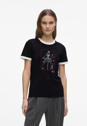 Γυναίκα που φοράει μαύρο T-shirt με λευκά περιθώρια και λευκό σκίτσο μόδας σχεδιασμένο με γραμμές, σε συνδυασμό με γκρι ριγέ παντελόνια.