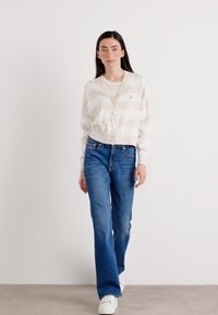 En kvinne går mot kameraet, ikledd en stripet cardigan, hvit skjorte og blå jeans, med hvite sneakers på en nøytral bakgrunn.
