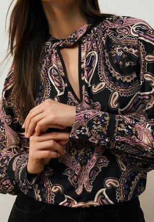 Femme portant un chemisier noir avec un motif paisley rose et beige et un col en forme de goutte, mains délicatement jointes à la taille.
