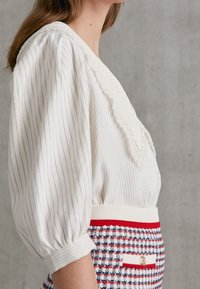 Vit blus med vertikala ränder, spetsdetalj längs halsringningen och puffärmar. Bärs med en röd, vit och blå rutmönstrad kjol.