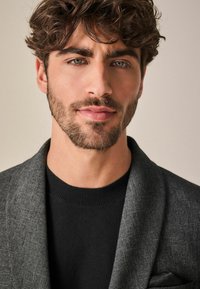 Blazer gris texturizado sobre un suéter negro, mostrando un corte entallado con solapa, llevado por un hombre con cabello despeinado y una barba ligera.