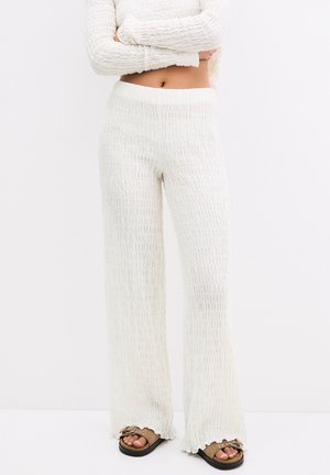 Femme portant un pantalon blanc texturé à taille haute et jambes larges, ainsi que des sandales plates marron à clous, se tenant devant un fond blanc uni.