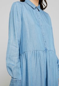 Femme portant une robe bleu clair à manches longues boutonnée avec une taille froncée et un col, vue des épaules jusqu'au milieu du torse.
