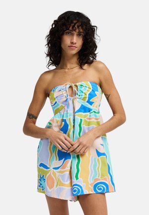 Vrouw met krullend donker haar, gekleed in een strapless, kleurrijke abstracte bloemenjurk, staand met samengevouwen handen ter hoogte van de taille tegen een effen achtergrond.