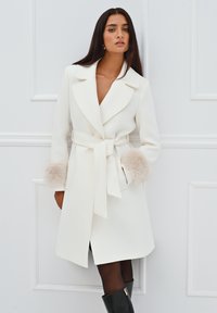Cappotto bianco con vita arricciata, ampi rever e polsini in pelliccia beige morbida. Taglio dritto con orlo a ginocchio e tasche laterali.
