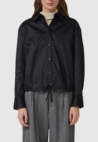 Kobieta w czarnej koszuli na guziki z subtelnym wzorem kropek, ze ściągaczem u dołu, nałożona na beżowy golf, w połączeniu z szarymi spodniami w cienkie prążki.