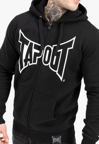 Czarna bluza z kapturem na zamek z dużym białym logo "TAPOUT", kieszeniami z przodu oraz ściągaczami przy mankietach. Wykonana z miękkiego materiału o gładkiej fakturze.