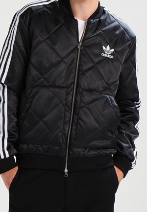Schwarze gesteppte Adidas-Jacke mit weißen Streifen an den Ärmeln und Logo auf der Brust, getragen und zugezippt von einer Person mit den Händen in den Taschen.