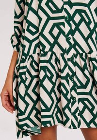 Grün und cremefarbenes gemustertes Kleid mit geometrischem Design, Knopfleiste und gerafftem Saum. Hergestellt aus leichtem Stoff mit glatter Textur.