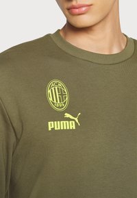 Sweat-shirt vert olive, col côtelé, avec logos jaunes AC Milan et Puma. Confectionné en tissu lisse avec une coupe ajustée.